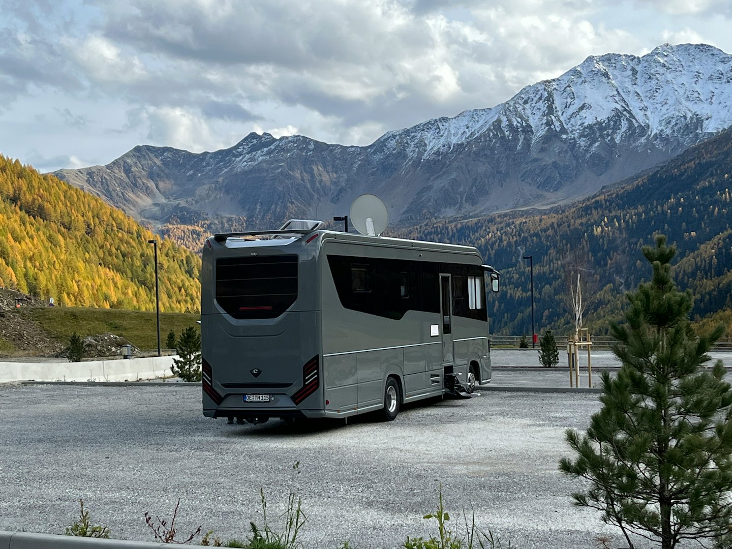 A grey motorhome parked in a gravel lot near snowy mountains.